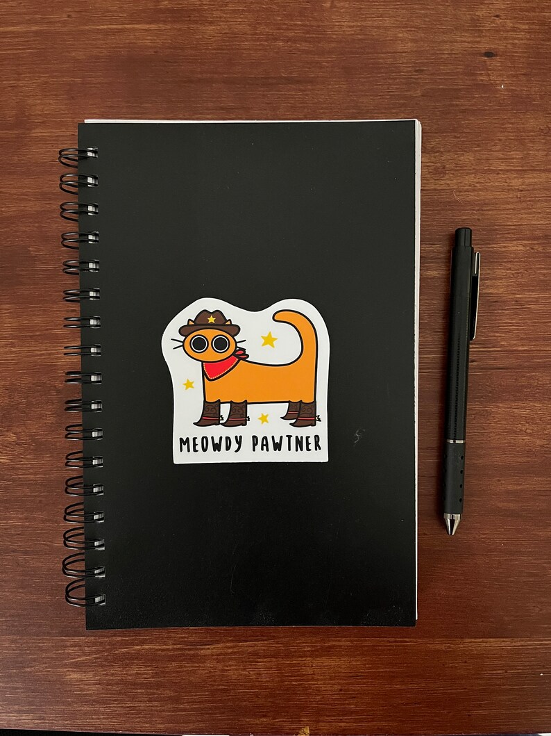 May include: A black spiral-bound notebook with a sticker of an orange cat wearing a cowboy hat and the text "MEOWDY PAWTNER." A black pen rests on a wooden surface next to the notebook.