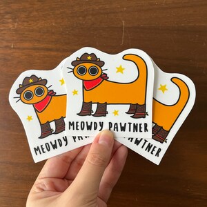 May include: Three stickers with a cartoon cat design. The cat is orange, wearing a cowboy hat, red neckerchief, and boots. The text "MEOWDY PAWTNER" is printed below the cat on each sticker.
