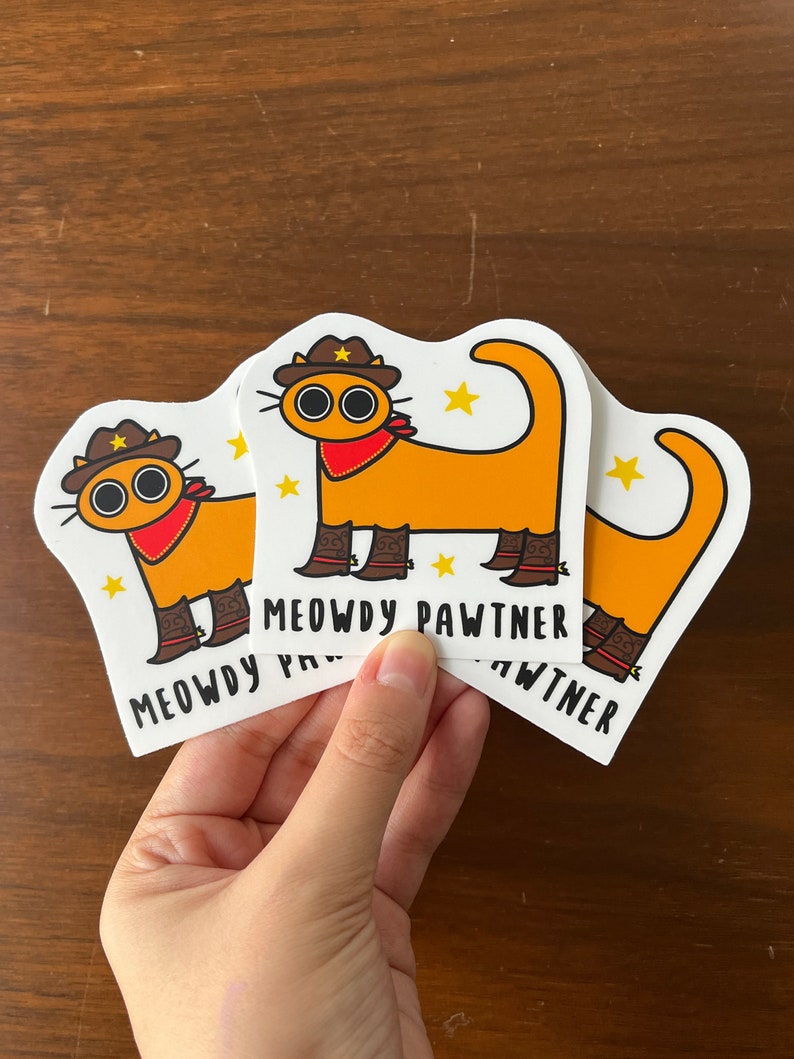 May include: Three stickers with a cartoon cat design. The cat is orange, wearing a cowboy hat, red neckerchief, and boots. The text "MEOWDY PAWTNER" is printed below the cat on each sticker.