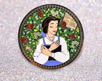 Op de afbeelding: Een ronde emaille pin met Belle uit Belle en het Beest. Belle is afgebeeld in een witte jurk met een blauwe buste, met een boek dicht tegen haar borst. Ze is omringd door rode rozen, groene bladeren en witte bloemen. De pin heeft een gouden rand met een decoratief patroon.