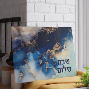 May include: A rectangular canvas print with an abstract design in shades of blue, white, and gold. The artwork features a fluid, marbled effect. The words "Shabbat Shalom" are written in black script on the lower right corner.