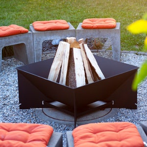 Könnte beinhalten: Ein schwarzer Metall-Feuerstelle mit gestapeltem Brennholz in der Mitte, umgeben von vier orangefarbenen Kissen auf Betonsockeln. Die Feuerstelle befindet sich auf einer Kiesfläche in einem Garten.