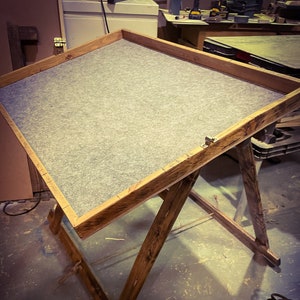 May include: A wooden table with a gray felt top and a rustic, distressed finish. The table is supported by two wooden legs that form an X shape.