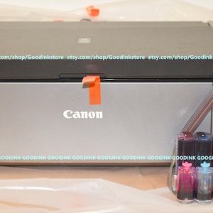May include: A silver Canon PRO-100 printer with a black top and a set of 10 ink cartridges in clear plastic containers. The cartridges are arranged in a row and are labeled with different colors.