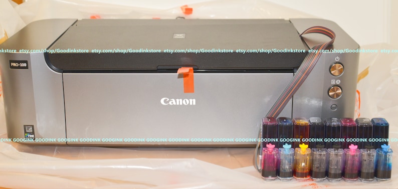 May include: A silver Canon PRO-100 printer with a black top and a set of 10 ink cartridges in clear plastic containers. The cartridges are arranged in a row and are labeled with different colors.