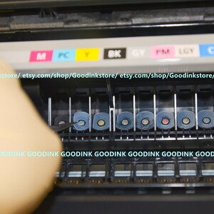 May include: Close-up of a printer ink cartridge replacement process. The printer has a row of eight ink cartridges, each with a different color: magenta, photo cyan, yellow, black, gray, photo magenta, light gray, and cyan. The cartridges are labeled with their corresponding colors.