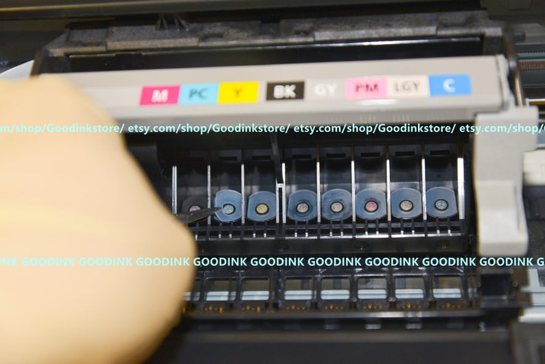May include: Close-up of a printer ink cartridge replacement process. The printer has a row of eight ink cartridges, each with a different color: magenta, photo cyan, yellow, black, gray, photo magenta, light gray, and cyan. The cartridges are labeled with their corresponding colors.