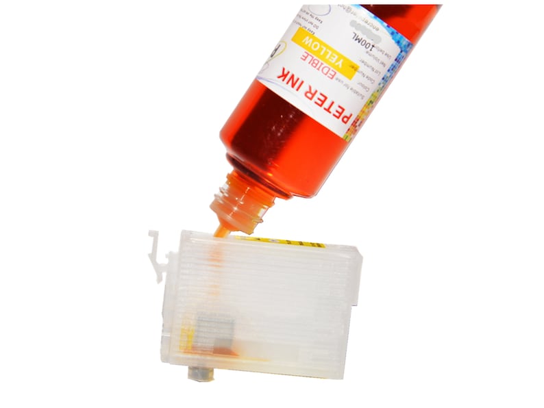 Op de afbeelding: Een transparante plastic inktcartridge wordt gevuld met gele eetbare inkt uit een fles met het label "PETER INK EDIBLE YELLOW 100ML".