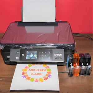 Puede incluir: Una impresora Epson XP-440 negra con una hoja de papel blanca en la bandeja de salida. El papel tiene una tarjeta de cumplea&ntilde;os colorida impresa en &eacute;l. Hay seis cartuchos de tinta en la parte delantera de la impresora.