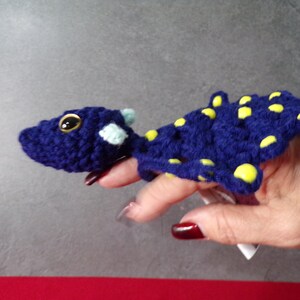 May include: A handmade, crocheted blue finger puppet shaped like a lizard or dragon, with yellow spots and a light blue accent. The puppet has large, round eyes and is held up by a finger.