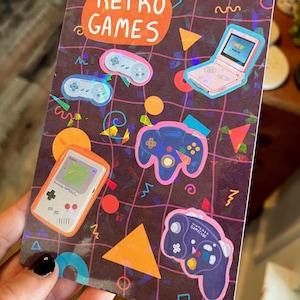May include: A rectangular sticker with a dark brown background featuring colourful illustrations of retro gaming consoles and controllers. The sticker has the words "Retro Games" in an orange bubble.