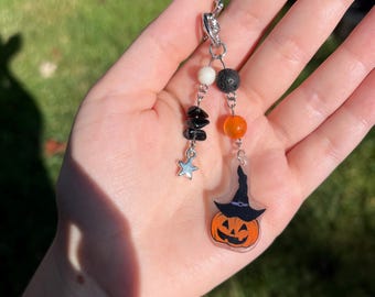 Puede incluir: Un llavero temático de Halloween con una calabaza sonriente con un sombrero de bruja. El llavero incluye cuentas negras, blancas y naranjas, un dije de estrella plateada y un cierre plateado. El fondo es un césped verde borroso.