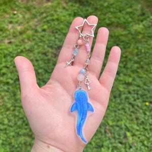 Puede incluir: Un amuleto de tiburón ballena azul con un amuleto de estrella plateada, cuentas rosas y azules y un amuleto de llave plateada colgando.