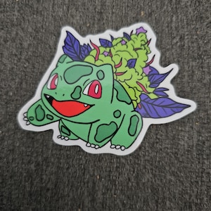 May include: A die-cut sticker featuring a green cartoon character with red eyes and a smiling red mouth. The character has green spots and is topped with a green and purple plant. The sticker is on a gray background.