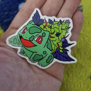 May include: A green sticker featuring a cartoon character with a smiling face, red eyes, and green spots. The character has a large green and purple plant on its back. The sticker is held in a hand.