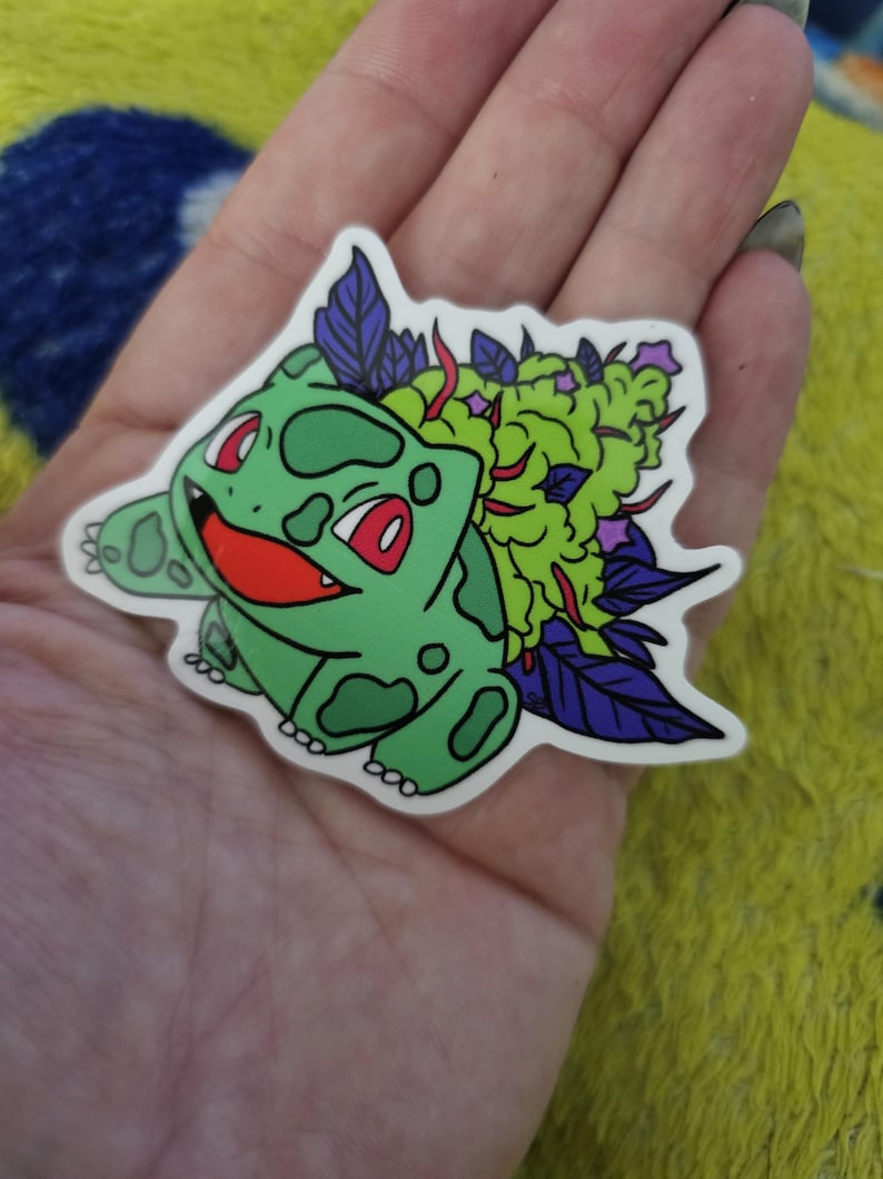 May include: A green sticker featuring a cartoon character with a smiling face, red eyes, and green spots. The character has a large green and purple plant on its back. The sticker is held in a hand.