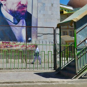 May include: A green metal gate with a person walking through it. A large poster of a man with a beard is on the wall behind the gate. The poster has text in a foreign language.