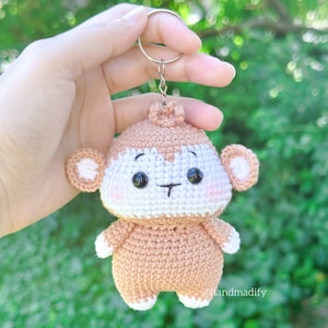 May include: A crocheted monkey keychain with brown fur, white facial features, and black eyes. The monkey is holding a key ring.