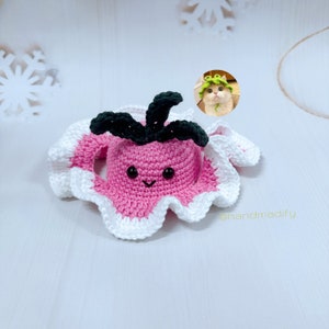 Peut inclure: Un chapeau en crochet rose et blanc en forme de champignon avec une tige en crochet verte et des yeux noirs. Le chapeau a un visage souriant et est posé sur une surface blanche. @handmadify