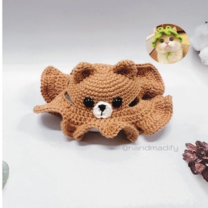 May include: A crocheted brown bear hat with a white nose and black eyes. The hat is shaped like a bear's head and has a floppy brim. The hat is sitting on a white surface.