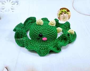 May include: Green crocheted amigurumi toy shaped like a dinosaur with yellow and white accents. The toy has a pink nose and black eyes.