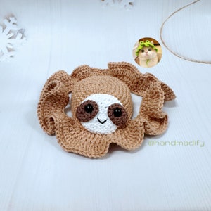 May include: A crocheted sloth toy with brown fur, white face, and black eyes. The sloth is wearing a green crocheted hat. The toy is on a white wooden surface. @handmadify