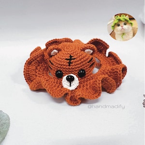 May include: A crocheted brown bear head with white eyes and a black nose, attached to a brown crocheted octopus-like body with ruffled tentacles. The bear head has a cross-shaped marking on its forehead. The image includes the text "@handmadify".
