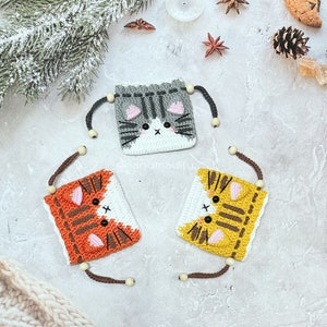 Puede incluir: Tres monederos de ganchillo con forma de gato en gris, naranja y amarillo, cada uno con cierre de cordón y cuentas de madera. Los monederos presentan detalles blancos y negros, con acentos rosas para las orejas y las mejillas. El fondo incluye elementos festivos.