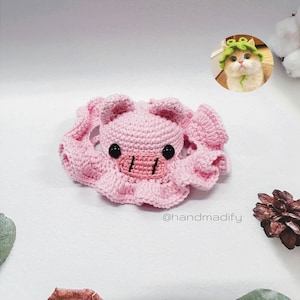 May include: A pink crocheted pig with black eyes and a pink snout. The pig is wearing a pink crocheted hat with a ruffled edge. The image includes the text "@handmadify".