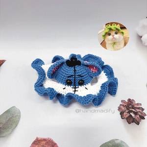 May include: A blue and white crocheted stuffed animal with a black nose and eyes. The stuffed animal is wearing a white crocheted dress with a ruffled hem. The stuffed animal is lying on a white surface. The image also includes a small, green crocheted hat on a white cat.