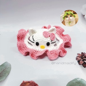 May include: A pink and white crocheted Hello Kitty hat with a white bow. The hat is on a white surface with a green leaf in the background.