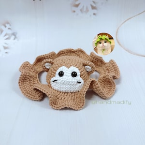 Może przedstawiać: Czapka małpy wykonana na szydełku w kolorze brązowym z białymi rysami twarzy i czarnymi oczami. Czapka leży na białym tle. W tle znajduje się małe zdjęcie kota w zielonym, liściastym kapeluszu. Tekst '@handmadify' znajduje się w prawym dolnym rogu obrazu.