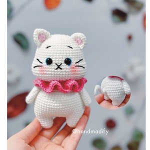 May include: White crocheted cat amigurumi toy with a pink ruffled collar and blue eyes. The cat is standing on a hand.