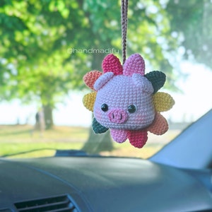 以下が含まれることがあります： 車の後部座席のミラーに、虹色の花模様のついた編みぐるみブタがぶら下がっています。ブタはピンクで、黒い目とピンクの鼻をしています。花は、さまざまな色の編みぐるみ花びらで作られています。