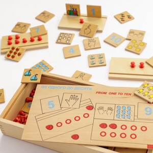Puede incluir: Juego educativo de madera con números, ilustraciones y clavijas rojas. El conjunto incluye una caja de madera con el texto "FROM ONE TO TEN" y fichas de números individuales. Las ilustraciones incluyen flores, barcos y gestos con las manos.