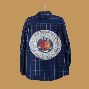 May include: A blue and white plaid flannel shirt with a circular design on the back. The design features a colorful face with a red and orange yin-yang symbol, surrounded by a circle of text that reads "SKYLAND TINGHAT MAZZA ONYANU OZZAIS".