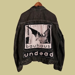 May include: A black denim jacket featuring a large rectangular patch on the back. The patch displays a black and white graphic of a bat-like figure with the text "bauhaus" above and the word "undead" below, all secured with visible stitching.