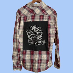 May include: Plaid button-down shirt in cream, red, and black. A black patch on the back features the words "NOTHING LESS THAN ABOLITION" above a graphic of a police van. The shirt has long sleeves and a collared neckline.