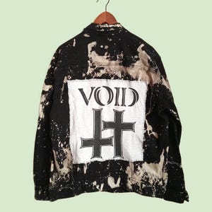 May include: A black denim jacket with a bleached splatter pattern and a large rectangular white fabric patch sewn onto the back. The patch features the word VOID in black lettering above two black cross symbols, one of which is inverted.