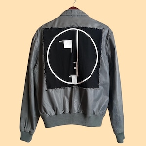 May include: Gray leather jacket with a black patch on the back featuring a white geometric design inside a circle. The jacket has a ribbed hem and cuffs, and is hanging on a wooden hanger.
