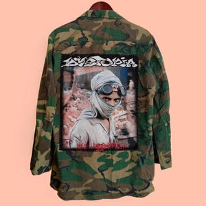 May include: Camouflage jacket with a large patch on the back featuring a person wearing goggles and a head covering. The patch has the word "AVISION" in white and the words "THE ARRESTED" in red.