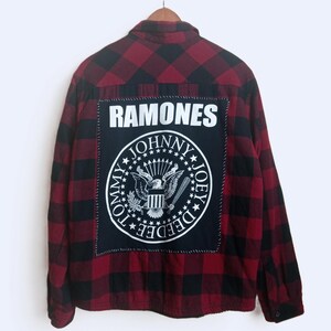 May include: Red and black plaid flannel shirt with a large black patch featuring the band name "RAMONES" and a circular emblem. The emblem includes an eagle, stars, and the names "Tommy", "Johnny", and "Joey".