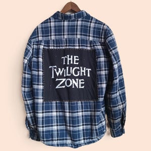 May include: Blue and white plaid flannel shirt with a black patch on the back. The patch features the text "THE TWILIGHT ZONE" in white lettering. The shirt is hanging on a wooden hanger.