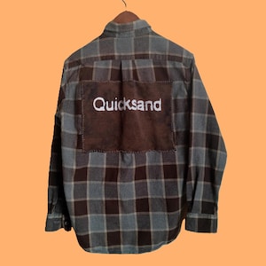 May include: A plaid flannel shirt in shades of brown and blue, with a brown patch on the back. The word "Quicksand" is embroidered in white on the patch. The shirt is hanging on a wooden hanger.