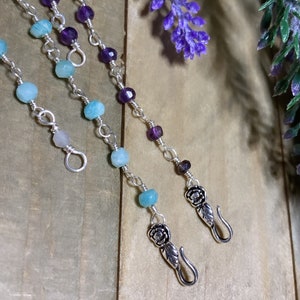May include: Two silver chains with gemstone beads and rose-shaped clasps. The chain on the left has light blue beads and the chain on the right has purple beads.