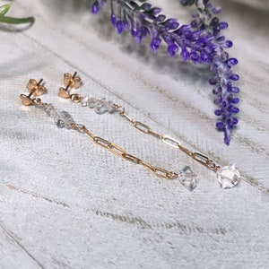 Puede incluir: Un par de pendientes de cadena dorados con detalles de cristal transparente facetado. Los pendientes se exhiben sobre una superficie de madera blanca, con ramitas de lavanda morada en el fondo. Los pendientes tienen cierre de poste.