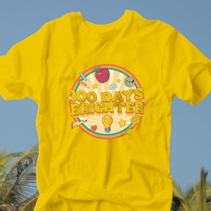 May include: A vibrant yellow t-shirt featuring the text "100 Days Brighter" in a retro design. The graphic includes an apple, pencil, and lightbulb. The shirt is made of a soft material and is ideal for celebrating a school milestone. The shirt is perfect for a school milestone.