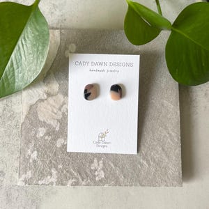 May include: A pair of small, square-shaped earrings with a pink, black, and white abstract pattern. The earrings are on a white card with the text "Cady Dawn Designs handmade jewelry".