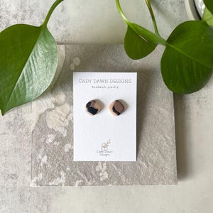 May include: A pair of small, square-shaped earrings with a pink, brown, and black abstract pattern. The earrings are displayed on a white card with the text "Cady Dawn Designs Handmade Jewelry".
