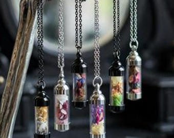 Puede incluir: Seis colgantes de frasco de vidrio cilíndrico rellenos de pétalos de flores secas de colores, suspendidos de finas cadenas de metal. Los collares cuelgan de un soporte de exhibición plateado con forma de mano esquelética.